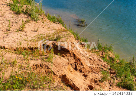 Steep sandy bank with sparse green grass bordering a calm turquoise lake on a sunny summer day, showcasing a picturesque natural erosion landscape near the water edge Steep sandy bank with sparse green grass bordering a calm turquoise lake on a sunny summer day, showcasing a picturesque natural erosion landscape near the water edge 142414338