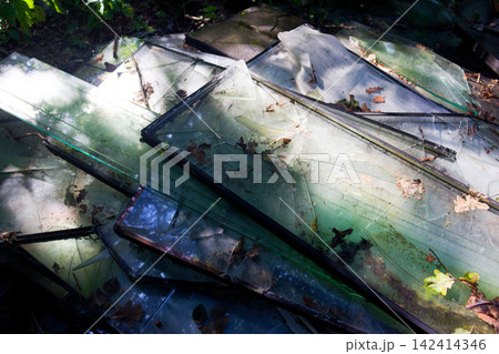 A pile of discarded broken window glass and shattered frames abandoned in the forest, creating a sharp and dangerous contrast against the soft natural woodland foliage A pile of discarded broken window glass and shattered frames abandoned in the forest, creating a sharp and dangerous contrast against the soft natural woodland foliage 142414346