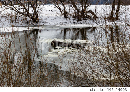 Drifting ice chunks float on a dark, nascent river, signaling early spring's thaw. Leafless trees on snow-covered banks reflect in the water, a stark, tranquil scene of seasonal transition 142414998