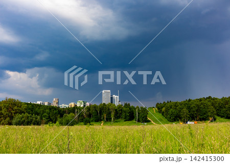 Dark ominous storm clouds gather over a vibrant green meadow and a lush forest with modern city buildings rising on the horizon under a dramatic moody sky before the heavy rain Dark ominous storm clouds gather over a vibrant green meadow and a lush forest with modern city buildings rising on the horizon under a dramatic moody sky before the heavy rain 142415300