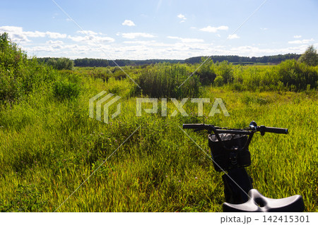 Close-up view of a folding fat tire e-bike handlebars parked in a vibrant green summer meadow, adventure travel concept, POV shot during a sunny outdoor ride through nature Close-up view of a folding fat tire e-bike handlebars parked in a vibrant green summer meadow, adventure travel concept, POV shot during a sunny outdoor ride through nature 142415301