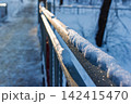 Glittering snow caps a bridge railing, bathed in golden hour light. Frosty metal shines brightly against a soft winter backdrop 142415470