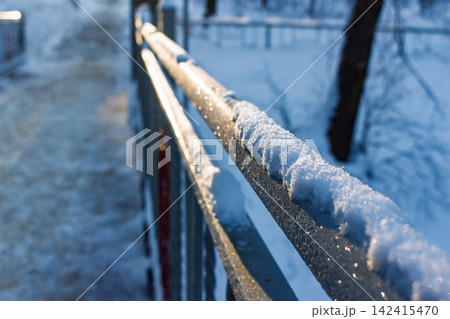 Glittering snow caps a bridge railing, bathed in golden hour light. Frosty metal shines brightly against a soft winter backdrop 142415470