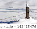 Gritty concrete fence post with barbed wire cuts across a vast snowy winter field. Sharp shadows stretch, emphasizing stark isolation and a defined perimeter 142415476
