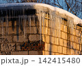 Glistening icicles cling to a snowy garage roof, bathed in warm golden sunset light. A frosty winter scene showcasing cold beauty and delicate ice formations 142415480