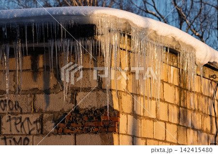 Glistening icicles cling to a snowy garage roof, bathed in warm golden sunset light. A frosty winter scene showcasing cold beauty and delicate ice formations Glistening icicles cling to a snowy garage roof, bathed in warm golden sunset light. A frosty winter scene showcasing cold beauty and delicate ice formations 142415480