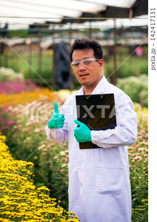 Researcher man giving thumbs up while holding clipboard in a flower nursery, wearing safety glasses, gloves and lab coat with confident positive expression Researcher man giving thumbs up while holding clipboard in a flower nursery, wearing safety glasses, gloves and lab coat with confident positive expression 142417131