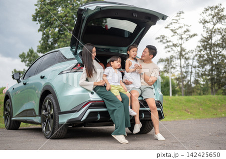 Asian family sharing a joyful conversation while sitting in an open SUV trunk in the green park. 142425650
