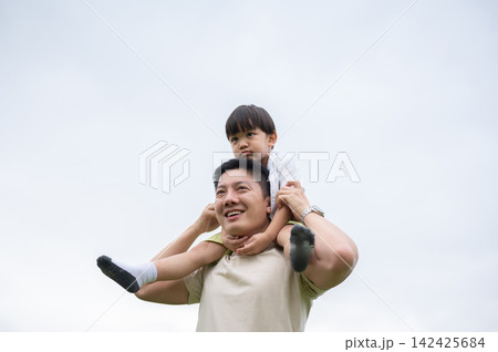 Asian father smiling while carrying a young kid son on his shoulders with a clear sky in background. 142425684