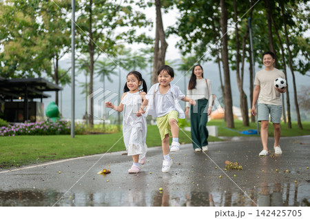 Asian family walking together through the park after rain with children running ahead on wet pathway 142425705