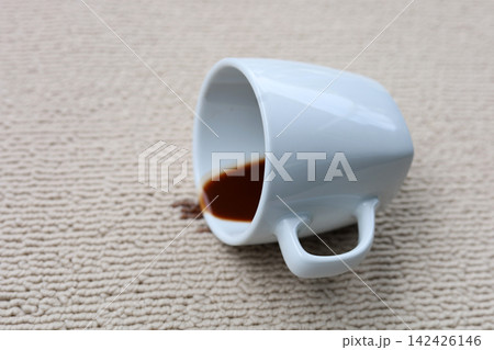 A white coffee cup has tipped onto its side, and dark coffee has spilled messily onto the carpet. A white coffee cup has tipped onto its side, and dark coffee has spilled messily onto the carpet. 142426146