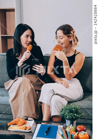 Two young female students using a laptop while sitting on a sofa at home. Student life concept with studying, teamwork, and online learning. 142428664