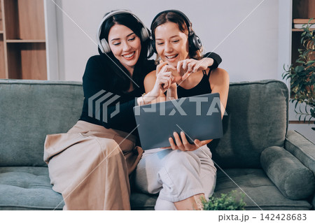Two young female students using a laptop while sitting on a sofa at home. Student life concept with studying, teamwork, and online learning. 142428683
