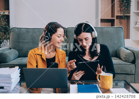Two young female students using a laptop while sitting on a sofa at home. Student life concept with studying, teamwork, and online learning. 142428708
