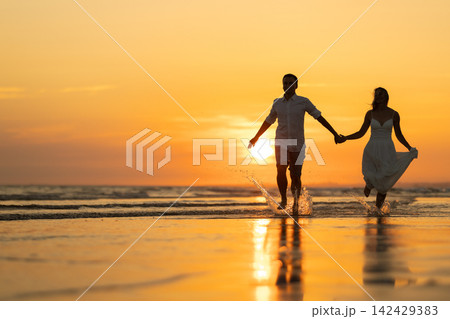 Couple enjoys time together on a beach during sunset while holding hands and playing in water Couple enjoys time together on a beach during sunset while holding hands and playing in water 142429383