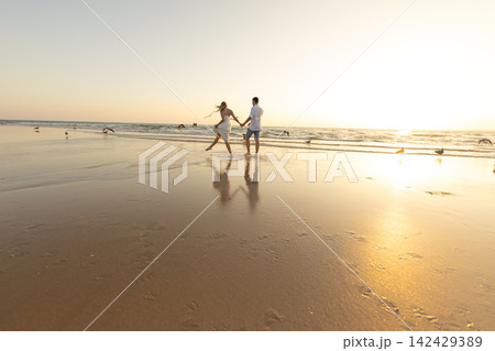 Couple walks hand in hand on the beach as the sun sets in the background 142429389