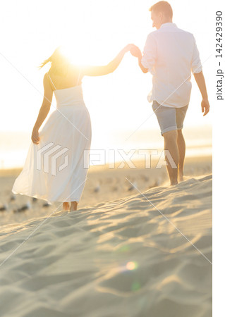 Young man and woman walk hand in hand on the beach during sunset with sun shining behind them Young man and woman walk hand in hand on the beach during sunset with sun shining behind them 142429390