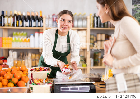 Woman sells and puts candies in bag for young woman, puts them on scale and weighs fruits Woman sells and puts candies in bag for young woman, puts them on scale and weighs fruits 142429786