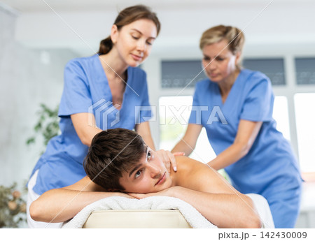 Female professional physiotherapist helps girl student to perform back massage to male client. Female professional physiotherapist helps girl student to perform back massage to male client. 142430009