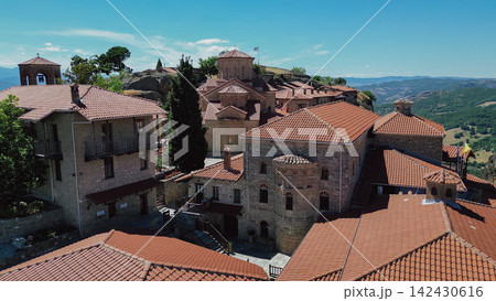 Scenic view of traditional stone buildings with red tiled roofs in Meteora, Greece, surrounded by lush greenery and distant mountains under a clear blue sky Scenic view of traditional stone buildings with red tiled roofs in Meteora, Greece, surrounded by lush greenery and distant mountains under a clear blue sky 142430616
