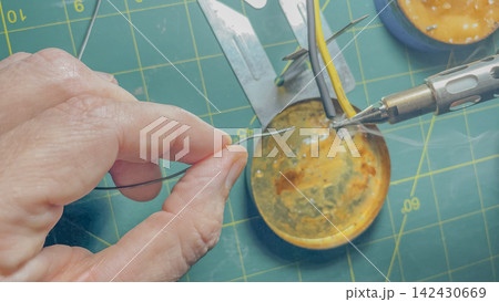 Close-up of a hand holding a thin wire while soldering electronic components on a green cutting mat with various tools and a soldering iron visible in the background Close-up of a hand holding a thin wire while soldering electronic components on a green cutting mat with various tools and a soldering iron visible in the background 142430669