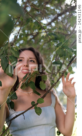 Smiling young woman with open eyes standing beneath leafy tree branches in sunlit park, holding foliage with calm relaxed expression, bright backlit greenery, soft portrait framing Smiling young woman with open eyes standing beneath leafy tree branches in sunlit park, holding foliage with calm relaxed expression, bright backlit greenery, soft portrait framing 142431146