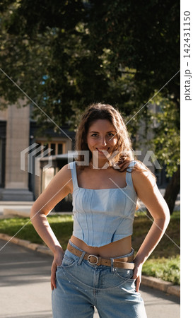 A portrait of a cheerful girl with long curly hair smiling at the camera. A close-up portrait of a friendly young woman in casual clothes against a blurred city background. Outdoors. 142431150