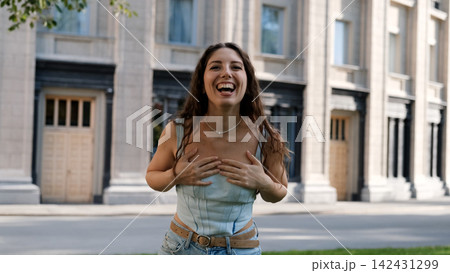 A portrait of a cheerful girl with long curly hair smiling at the camera. A close-up portrait of a friendly young woman in casual clothes against a blurred city background. Outdoors. A portrait of a cheerful girl with long curly hair smiling at the camera. A close-up portrait of a friendly young woman in casual clothes against a blurred city background. Outdoors. 142431299