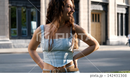 A portrait of a cheerful girl with long curly hair smiling at the camera. A close-up portrait of a friendly young woman in casual clothes against a blurred city background. Outdoors. A portrait of a cheerful girl with long curly hair smiling at the camera. A close-up portrait of a friendly young woman in casual clothes against a blurred city background. Outdoors. 142431308