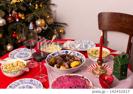 A table set for a Christmas and New Year celebration displays various dishes like salad, meat, and grains. A red candle, wine glass, and colorful decorations add to the festive atmosphere 142432817