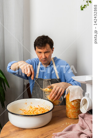 A 53-year-old man works in his kitchen fermenting cabbage and carrots. He carefully adds the mixture from a large bowl into a jar, showing concentration 142432868
