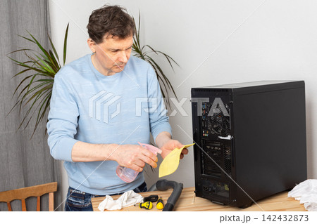 A 53-year-old man removes dust from a computer system unit while standing at a wooden table. He holds a yellow sticky note and spray cleaner in his hands 142432873