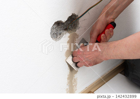 A person smooths plaster onto a white apartment wall, carefully applying it around a hole where an electrical outlet will be installed A person smooths plaster onto a white apartment wall, carefully applying it around a hole where an electrical outlet will be installed 142432899
