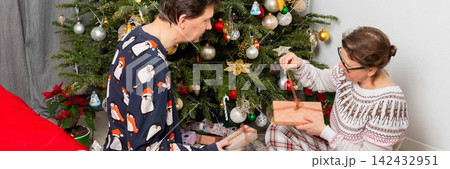 A couple sits near a Christmas tree on Christmas Day. They are exchanging gifts and enjoying the holiday spirit. The room has decorations and a festive atmosphere A couple sits near a Christmas tree on Christmas Day. They are exchanging gifts and enjoying the holiday spirit. The room has decorations and a festive atmosphere 142432951