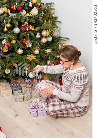 A woman aged 52 sits on the floor next to a decorated Christmas tree. She opens gifts on Christmas Day while enjoying the holiday with family members in the background A woman aged 52 sits on the floor next to a decorated Christmas tree. She opens gifts on Christmas Day while enjoying the holiday with family members in the background 142432953