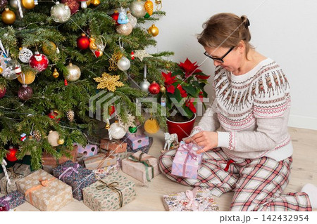 A woman aged 52 sits on the floor next to a decorated Christmas tree. She opens gifts on Christmas Day while enjoying the holiday with family members in the background 142432954