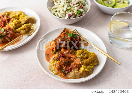 Plates hold amaranth, millet, and quinoa porridge served with baked white hake in tomato sauce. Fresh salad is beside the porridge. A bowl of kiwi sits nearby for dessert 142433408