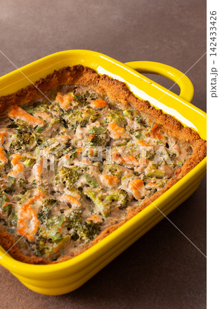 A gluten-free pie is placed in a bright yellow baking dish. It contains pieces of red fish and broccoli mixed with other ingredients. This dish is suitable for breakfast or lunch 142433426