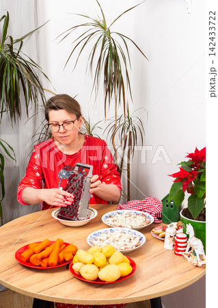 A woman grates beets for the Shuba salad on a Christmas table. Various ingredients are laid out, including herring, potatoes, eggs, and carrots, creating a festive atmosphere for the celebration A woman grates beets for the Shuba salad on a Christmas table. Various ingredients are laid out, including herring, potatoes, eggs, and carrots, creating a festive atmosphere for the celebration 142433722