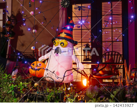 A white inflatable ghost wearing a striped witch hat stands beside a pumpkin on a residential porch. Purple lights, spiders, webs, and eyeballs create a colorful Halloween display at night. 142433927