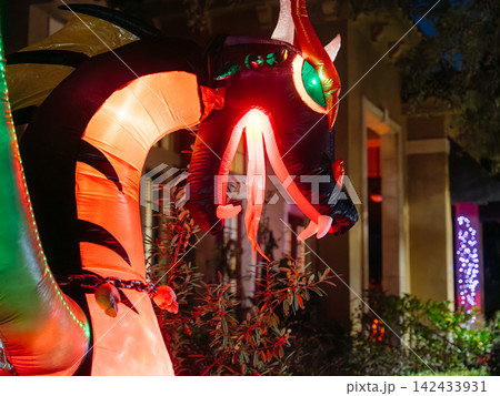 A giant red and black dragon inflatable glows outside a residential building. Bright horns, fangs, wings, and green eyes create a dramatic Halloween display. 142433931