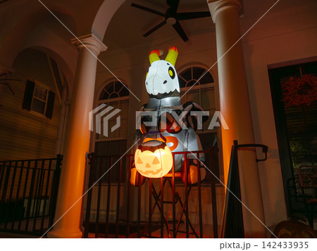 A large inflatable skeleton zebra holds a glowing pumpkin lantern on a decorated house porch. Orange lights illuminate the columns and create a spooky Halloween atmosphere at night. 142433935