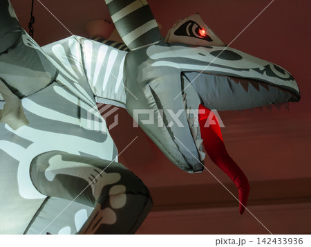 A glowing inflatable skeleton dragon with bright red eyes decorates a front porch for Halloween. The dramatic close-up highlights the creature's fierce appearance. A glowing inflatable skeleton dragon with bright red eyes decorates a front porch for Halloween. The dramatic close-up highlights the creature's fierce appearance. 142433936