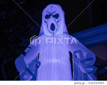 A glowing ghost inflatable with a haunting face stands outside during Halloween evening. Blue and purple lighting adds a dramatic spooky atmosphere. A glowing ghost inflatable with a haunting face stands outside during Halloween evening. Blue and purple lighting adds a dramatic spooky atmosphere. 142433947