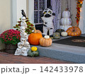 A charming Halloween porch showcases stacked skulls, pumpkins, colorful chrysanthemums, and a playful mummy cat decoration. The festive display celebrates the autumn season with creative outdoor decor 142433978