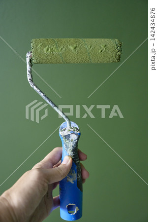 Hand holding paint roller covered with green paint in front of freshly painted wall Hand holding paint roller covered with green paint in front of freshly painted wall 142434876