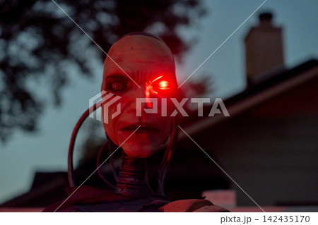 Frontal view of a cyborg head staring with a bright red eye at night. Metallic neck parts and cables create a creepy Halloween sci-fi scene. Frontal view of a cyborg head staring with a bright red eye at night. Metallic neck parts and cables create a creepy Halloween sci-fi scene. 142435170