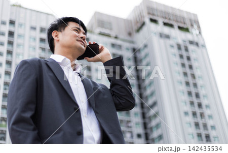 Confident Asian businessman talking on smartphone outdoors with modern office building blurred background. Professional corporate executive calling client during career commute in city center urban Confident Asian businessman talking on smartphone outdoors with modern office building blurred background. Professional corporate executive calling client during career commute in city center urban 142435534