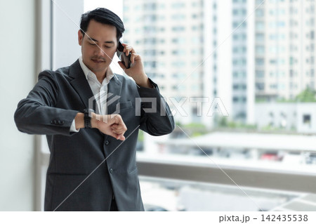 Busy Asian businessman checking wrist watch while talking on smartphone near office window. Professional corporate executive managing time during urgent mobile phone call in modern high rise building Busy Asian businessman checking wrist watch while talking on smartphone near office window. Professional corporate executive managing time during urgent mobile phone call in modern high rise building 142435538