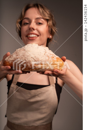 Smiling woman baker holding fresh artisan bread from bakery wearing a café themed apron showing homemade organic food Smiling woman baker holding fresh artisan bread from bakery wearing a café themed apron showing homemade organic food 142436154